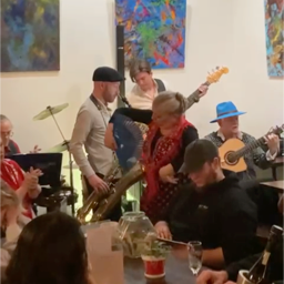 Aurora Reyes dances Guajiras con Guajeo in front of musicians. Aurora with fan amidst a turn, wearing black costume with red mantoncillo, with black fan, maroon border. Musicians Left to Right upstage Rex Benincasa modified cajon drum kit, Aron Caceres electric bass, Basilio Georges seated with guitar, singing, wearing bright blue Veracruz brimmed hat, Danny Rivera downstage left of Aurora standing playing baritone saxophone, facing right.