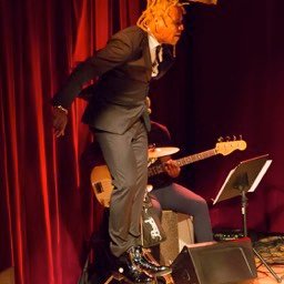 Omar Edwards appears to levitate vertically against stage right curtain wing in front of bass player. Omar, blonde braids, in black suit, white shirt black tie, black ankle high tap boots.